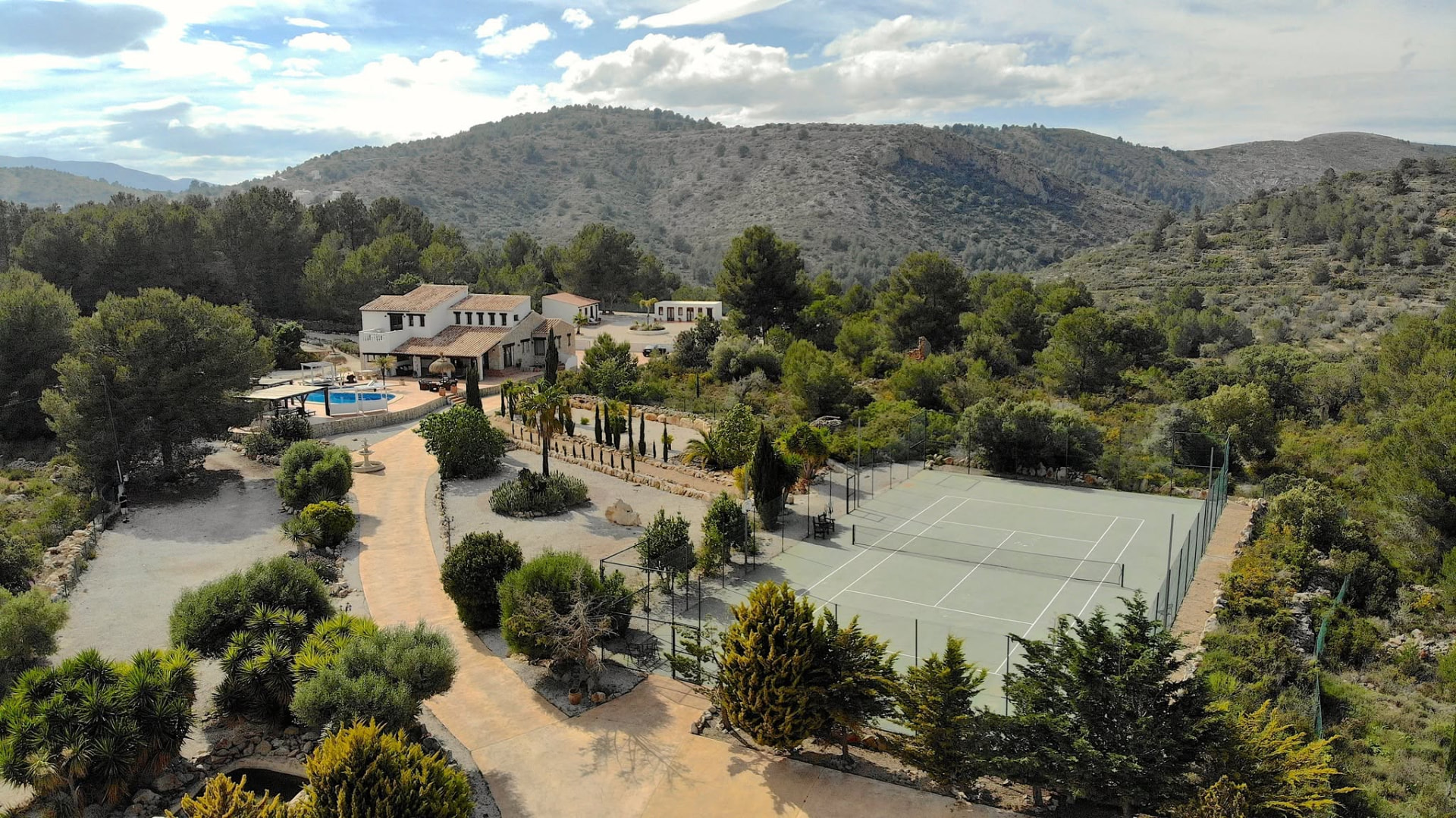 Aerial view of a white Spanish villa with swimming pool and tennis court set among trees and hills